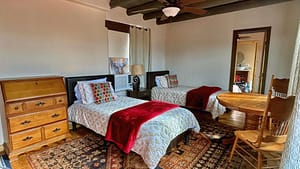 Interior of a cozy loft room at The Chi Center designed for relaxation and Wisdom Healing Qigong practice. The room features two single beds with white and patterned bedding, each accented with a red throw blanket, a traditional wooden dresser, and a small dining table with chairs. Richly patterned rugs cover the floor, enhancing the warm and inviting atmosphere, perfect for reflection and tranquility.