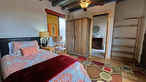 A cozy and inviting loft room at The Chi Center, ideal for relaxation and Wisdom Healing Qigong practice. This room features a comfortable bed with an ornate orange and white patterned bedspread and a dark headboard. Accents include a rustic wooden wardrobe, a wooden chair beside a round side table with a large lamp, and colorful abstract art on the walls. Visible in the background is the entrance to an en-suite bathroom, enhancing the sense of privacy and comfort.
