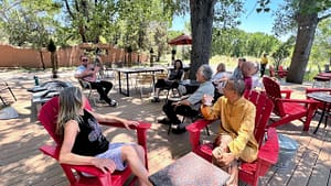 A relaxed gathering of people seated outdoors under the shade of a large tree, enjoying conversation and refreshments on a sunny day. Red chairs and tables add warmth to the serene natural setting, and participants appear engaged and content. This tranquil moment captures a sense of community, connection, and gratitude, reflecting the peaceful spirit of a Magical Sunday.