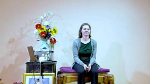 Rebecca Rule shares her testimonials at a fall retreat, seated with a gentle and reflective expression. Holding a microphone, she sits beside a vibrant floral arrangement and spiritual icons, creating an inviting atmosphere for personal reflection, community connection, and shared healing experiences.