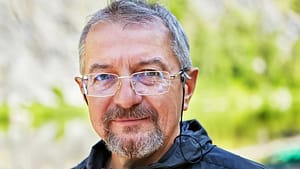 A middle-aged man with glasses and a short beard smiles softly while standing outdoors, with a blurred green natural background.