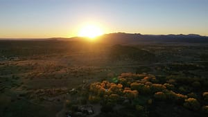 Aerial view of sunrise over the high desert landscape of Galisteo, New Mexico, with golden light illuminating the Sangre de Cristo mountains and casting a warm glow on cottonwood trees surrounding The Southwest Sanctuary retreat center. The scene highlights natural serenity, expansive skies, and the peaceful, healing energy of this sacred land.