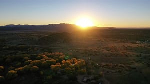Aerial view of sunrise over the high desert landscape of Galisteo, New Mexico, with golden light illuminating the Sangre de Cristo mountains and casting a warm glow on cottonwood trees surrounding The Southwest Sanctuary retreat center. The scene highlights natural serenity, expansive skies, and the peaceful, healing energy of this sacred land.