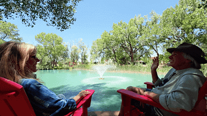 Two people sit in bright red chairs by a turquoise pond with a central fountain, surrounded by tall green trees under a clear blue sky at The Southwest Sanctuary.