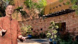 Master Mingtong Gu stands smiling in front of The Southwest Sanctuary in Galisteo, New Mexico, wearing a traditional red Chinese jacket. The scene features natural stone walls, adobe architecture, blue ceramic pots, and lush desert landscaping beneath cottonwood trees—offering a serene and welcoming space for healing retreats and spiritual renewal.