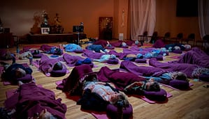 Participants lying on mats with blankets and pillows in a serene Kiva Healing session, led by an instructor in a softly lit room with spiritual decor, promoting relaxation and holistic well-being.