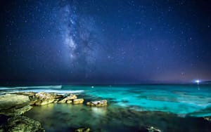 A serene night sky, speckled with countless stars, featuring the Milky Way stretching above a tranquil, glowing turquoise sea. The foreground shows rugged rocky outcrops at the water's edge, enhancing the peaceful yet awe-inspiring ambiance of the scene, reflecting the meditative and cosmic connection emphasized in Wisdom Healing Qigong practice.