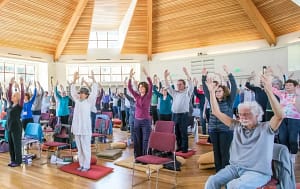 Large group of people practicing Qigong indoors with hands raised, in a bright and spacious room with natural light and a wooden ceiling."