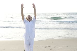 A man standing on a beach with his arms in the air practicing Qigong.