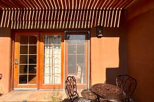 Patio view of a serene adobe-style building with wooden French doors, a glass screen door, and a wrought iron table and chairs under a pergola casting shadow patterns.