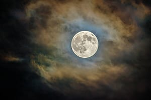 A full moon radiates a luminous, serene glow amidst swirling clouds, symbolizing tranquility and the reflective nature of a full moon Wisdom Healing Qigong practice. The moon's detailed craters and bright surface are stark against the dark, moody backdrop of the night sky, inviting a sense of calm and centeredness ideal for meditation.