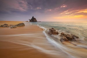 Golden sandy beach at sunset with gentle waves flowing over rocks, vibrant colors in the sky, and a small island in the distance, evoking a sense of tranquility and natural beauty.