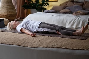 Senior woman practicing a relaxing yoga pose, lying on her back in Savasana on a mat in a cozy, softly lit bedroom with natural decor and cushions.