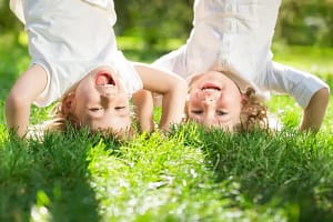 Two children playfully hanging upside down on a grassy lawn, laughing and enjoying a sunny day.