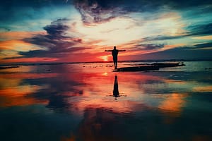 A silhouette of an individual at the shoreline, with arms extended, reflecting Life against a breathtaking sunset and tranquil waters.