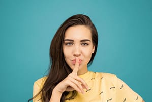 A young individual with long brown hair, wearing a yellow shirt with repeated "STOP" text, poses against a turquoise background. They place a finger to their lips in a "shushing" gesture, looking directly at the viewer with a playful expression.