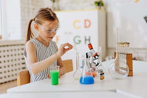 Young girl wearing safety goggles conducting a science experiment with colorful liquids in test tubes and beakers, fostering curiosity and hands-on learning in a classroom setting.
