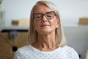 Calm mature woman with glasses and closed eyes, practicing mindfulness or relaxation indoors in a peaceful setting.