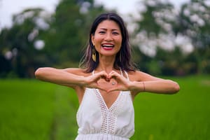 Joyful woman in a white dress standing in a lush green field, forming a heart shape with her hands.