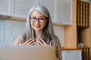 Smiling mature woman with gray hair and glasses wearing earphones, engaged in an online video call from her kitchen.