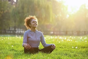 Young woman sitting cross-legged on grass in a serene park, smiling and enjoying the warm sunlight with a peaceful expression.
