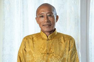Portrait of Master Mingtong Gu smiling gently, wearing glasses and dressed in a traditional golden yellow robe with intricate patterns, standing in front of a softly lit lace-curtained window.
