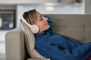 A woman relaxing on a couch with headphones on, eyes closed, and a peaceful expression, immersed in a listening experience. She is wrapped in a cozy blue sweater, symbolizing comfort and tranquility, used for Spring 2024 Retreats - Kiva Healing Livestreams.