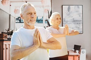 Smiling man and woman practicing Wisdom Healing Qigong with palms together in a prayer-like pose, radiating joy, inner peace, and mindfulness in a serene space