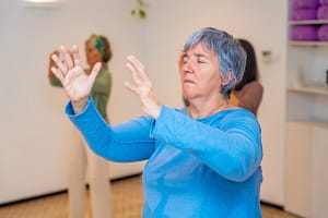 Woman practicing Wisdom Healing Qigong in a calm indoor space, focusing on gentle hand movements and energy flow, with others practicing in the background.
