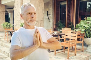 Smiling man practicing Wisdom Healing Qigong with palms together in a prayer-like pose, radiating joy, inner peace, and mindfulness in a serene space
