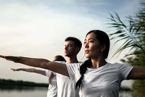 A group of focused individuals practicing a mindful stance by a serene lake, embodying the principles of Living Wisdom Mastery. Their open, extended arms represent a journey toward healing, liberation, and contribution, as they connect with nature and cultivate inner balance.