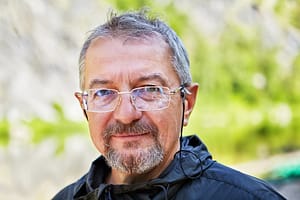A middle-aged man with glasses and a short beard smiles softly while standing outdoors, with a blurred green natural background.
