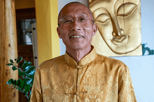 Master Mingtong Gu smiling warmly, wearing a golden silk tunic with intricate patterns, standing in a serene indoor setting with a golden Buddha artwork in the background. Perfect for themes of mindfulness, spiritual teachings, and holistic wellness.