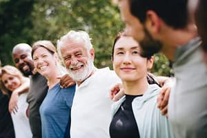 A diverse group of people standing outdoors in a line with their arms around each other's shoulders, smiling warmly and looking happy in a natural setting.