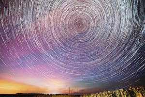 A mesmerizing long-exposure shot of star trails forming circular patterns in the night sky, with a colorful gradient of purple, pink, and orange hues near the horizon.