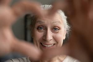 A cheerful older woman with gray hair smiles warmly while framing her face with her hands, creating a heart shape in the foreground.