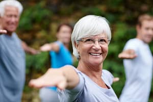 A joyful senior woman with white hair practicing outdoor group yoga or fitness, smiling confidently at the camera while stretching her arms forward. A diverse group of individuals participates in the background.