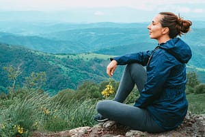 A woman sits peacefully on a rocky hillside, holding a small bouquet of yellow wildflowers, gazing out at a panoramic view of lush green hills and valleys under a cloudy sky. She wears a blue jacket and leggings, embodying tranquility and connection with nature.