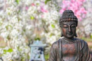 Bronze Buddha statue in peaceful meditation pose set against a vibrant spring garden with blooming white and pink flowers, symbolizing mindfulness, inner peace, and spiritual awakening in a natural setting.