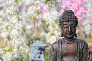 Bronze Buddha statue in peaceful meditation pose set against a vibrant spring garden with blooming white and pink flowers, symbolizing mindfulness, inner peace, and spiritual awakening in a natural setting.