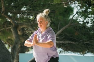 A woman with gray hair tied up in a bun, dressed in a lavender hoodie, is practicing a standing meditative pose outdoors.