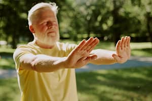 An elderly man with a white beard practicing Qigong in a sunlit park, focusing on mindful movement and energy flow for health, balance, and well-being.