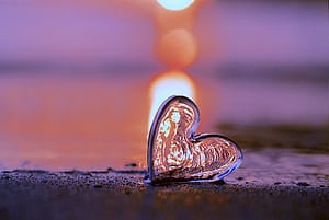 Glass heart on a sandy surface reflecting warm light at sunset, with a glowing background creating a peaceful and romantic atmosphere.