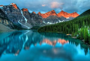 A serene mountain landscape with snow-capped peaks illuminated by the warm glow of sunset, reflected in a pristine turquoise lake surrounded by dense evergreen forests. Ideal for themes of natural beauty, tranquility, and adventure.