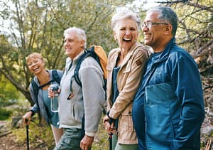 A joyful group of older adults enjoying a hike together in a lush, wooded area. They are dressed casually with backpacks, some holding water bottles, and are engaged in shared laughter, exuding warmth, friendship, and vitality. This image reflects the essence of 'Embrace Your Golden Years,' symbolizing the benefits of staying active, fostering connections, and finding joy in conscious aging through practices like Qigong.