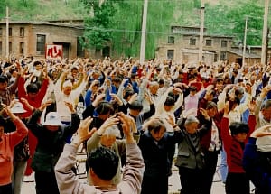 Large group of people practicing Qigong outdoors in a community gathering, performing synchronized movements with arms raised, surrounded by greenery and traditional buildings.