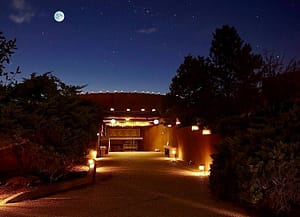 Night view of The Chi Center's Kiva illuminated by soft pathway lights, with dense trees surrounding the area under a star-studded sky and a luminous full moon.