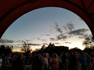 A view from inside a tent or dome, looking out at a gathering of people during sunset. The silhouettes of attendees, trees, and buildings are visible against a backdrop of a colorful evening sky with scattered clouds, creating a peaceful and social atmosphere