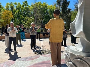 A group of individuals practicing mindfulness and meditation under the guidance of Master Mingtong Gu in a golden robe, gathered outdoors near a serene statue on a sunny day with lush greenery in the background.