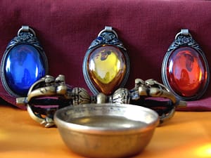 Three vibrant gemstone pendants in detailed silver settings—blue, yellow, and red—displayed alongside an ornate metal bowl, against a deep burgundy background, evoking a sense of spiritual energy and mysticism.